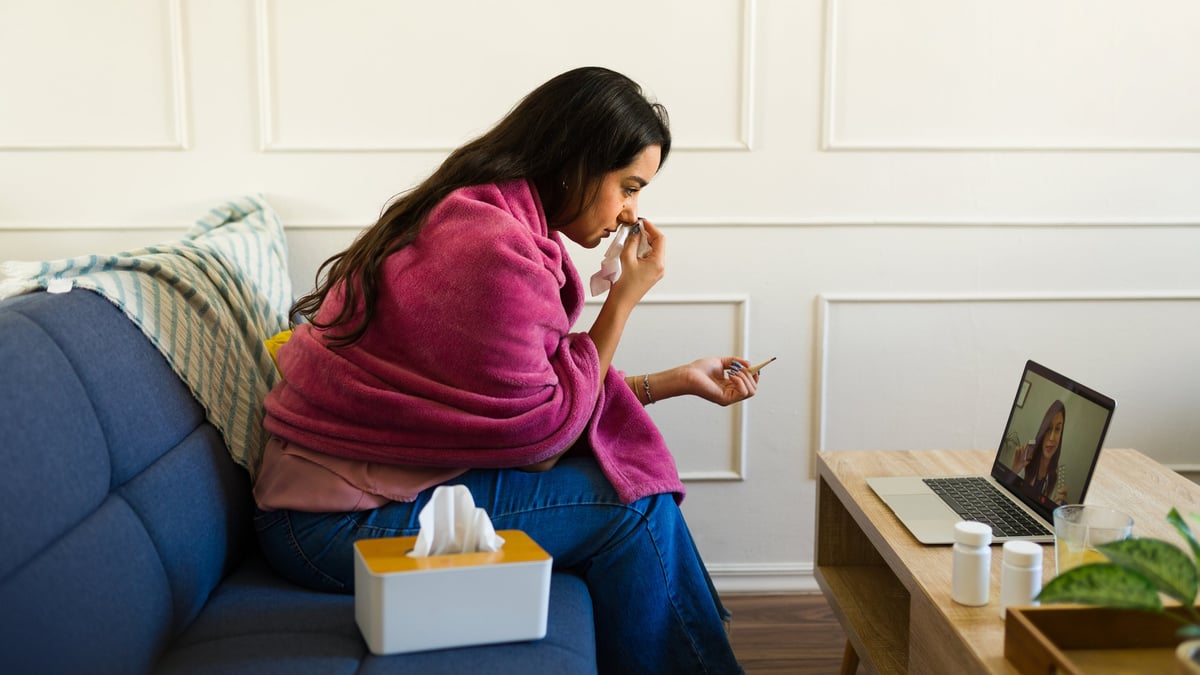 Sick young woman wrapped in a blanket is sitting on the sofa blowing her nose while having a video call with a doctor