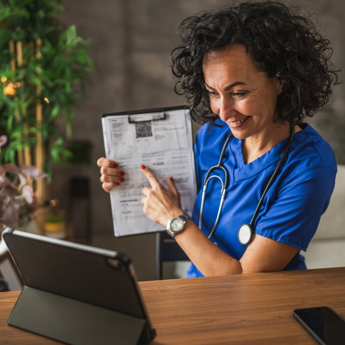 A medical professional, wear a stethoscope and blue scrubs, sit at a desk conducting a virtual consultation use a tablet