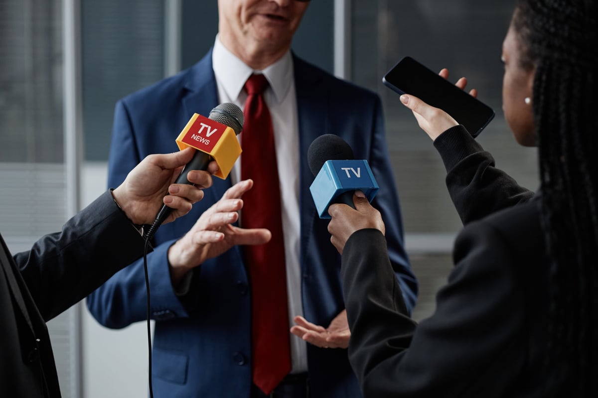 Cropped shot of middle aged Caucasian man in suit gesturing while giving interview by group to journalists holding microphones and smartphone in office room